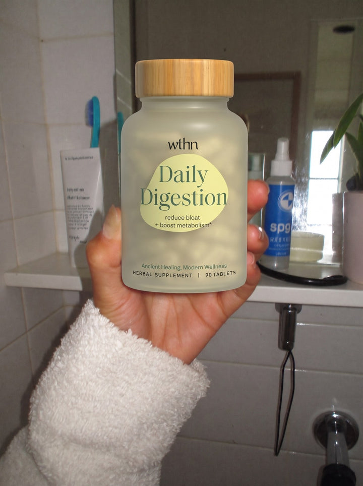 A hand in a bathrobe sleeve holding a bottle of Daily Digestion in the bathroom