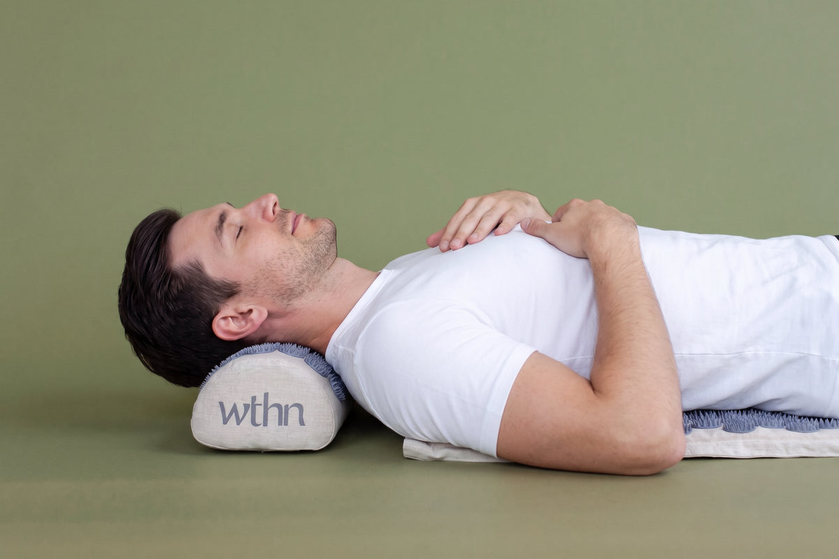 A man in a white T-shirt lying on the WTHN acupressure mat, his head on the wthn pillow, eyes closed