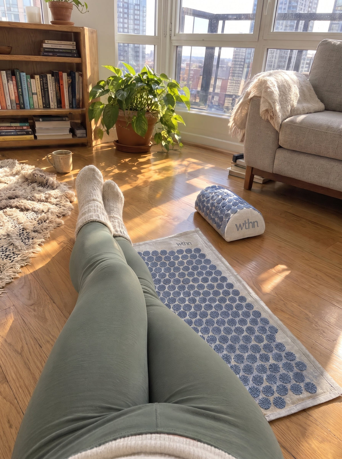 Legs in wool socks stretched out next to the WTHN acupressure mat and pillow on a sunny wooden floor