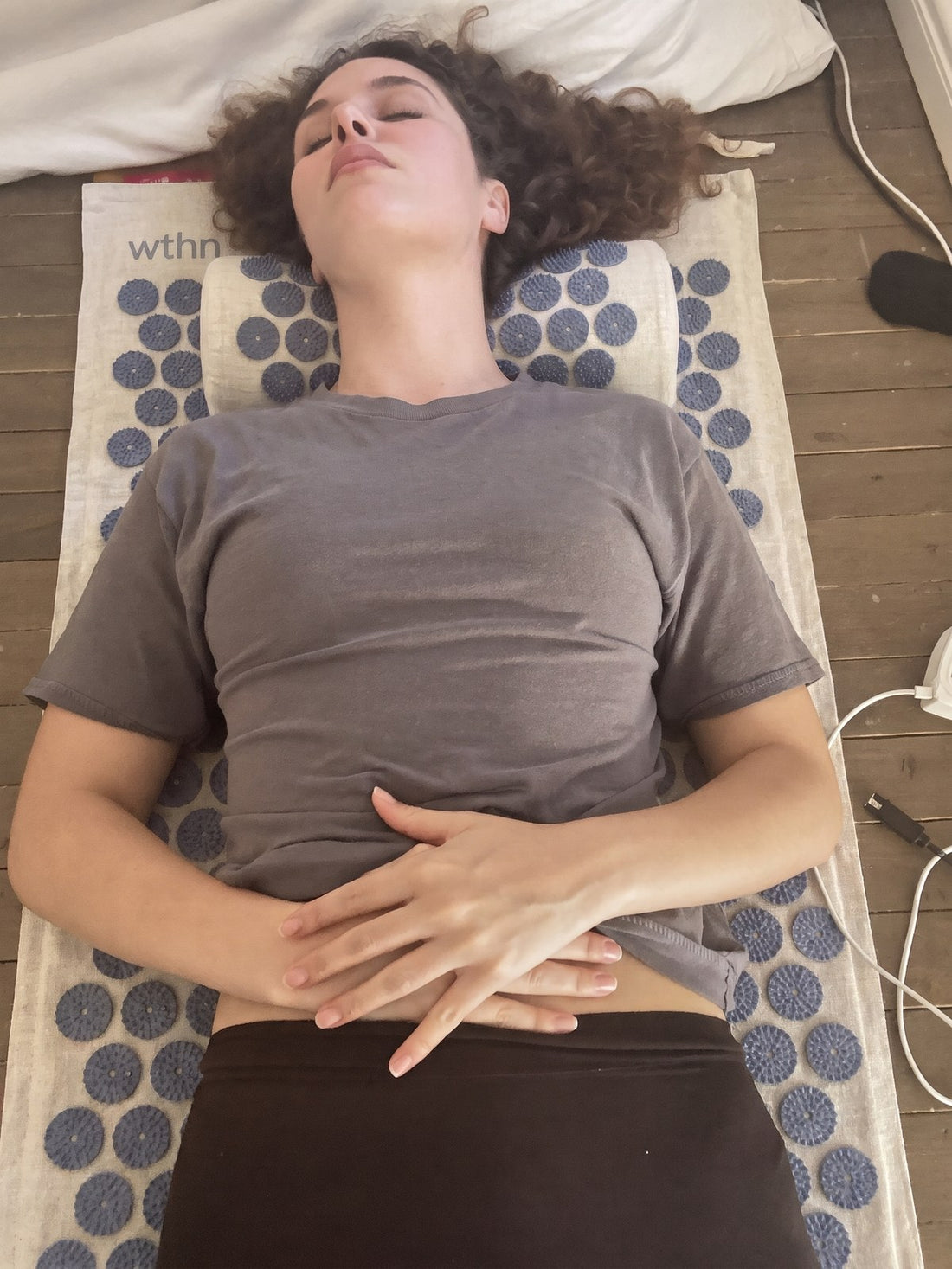 A woman resting on the WTHN acupressure mat on a bedroom floor in morning light
