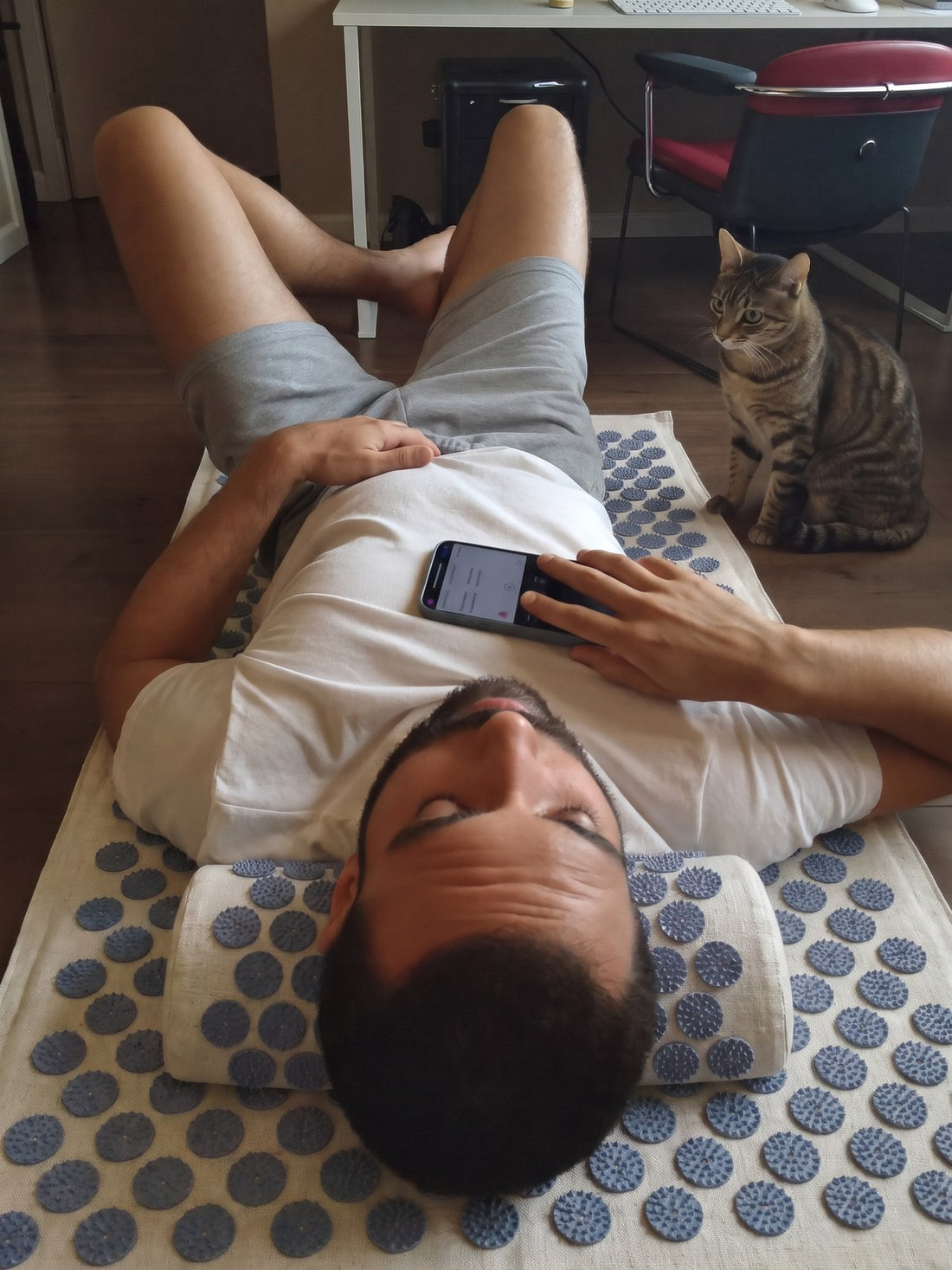 A man lying on the WTHN acupressure mat in a home office, a cat sitting beside him